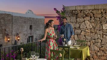 Una terraza con muros de piedra, una mesa lista para cenar y un atardecer de fondo.