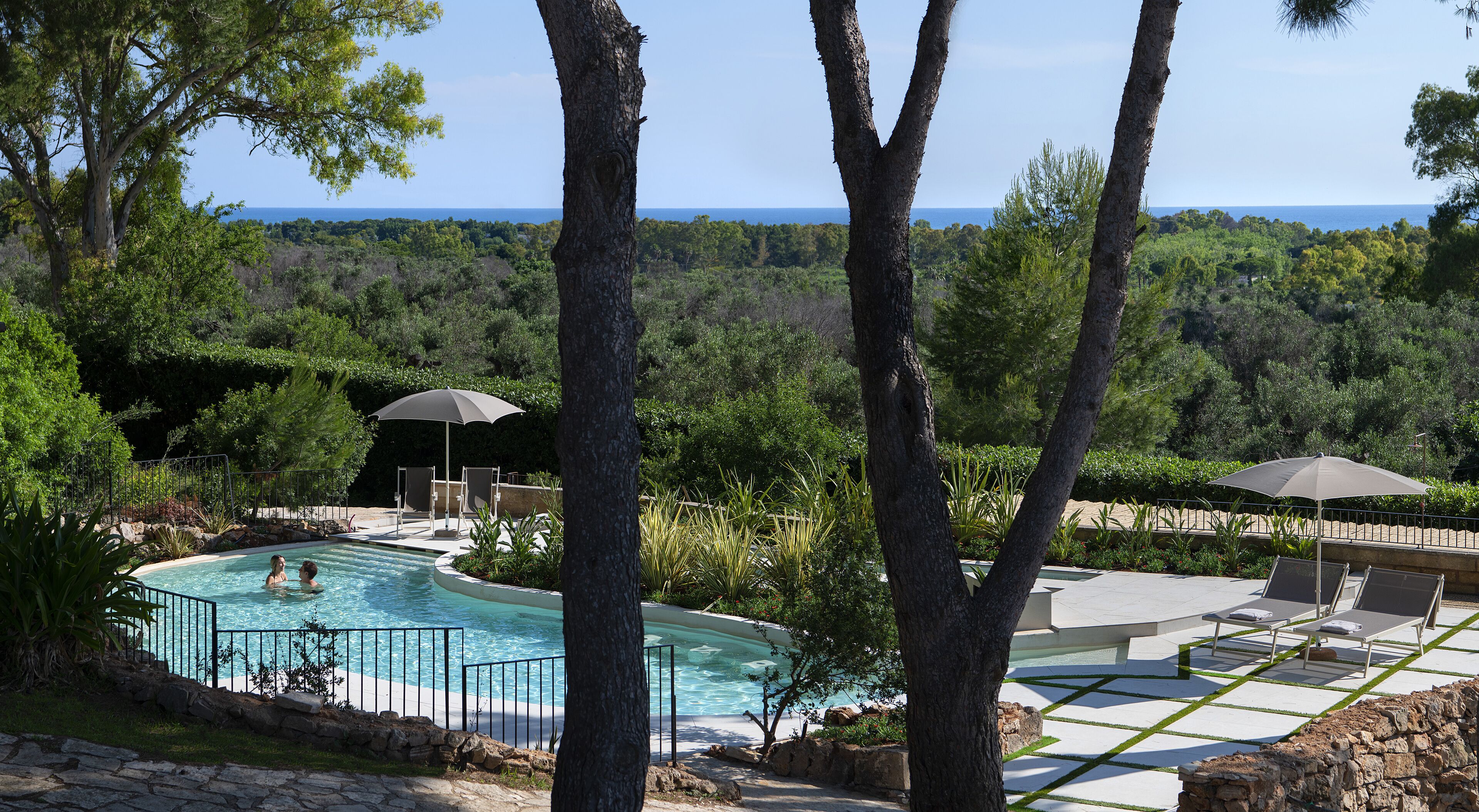 Area piscina con vista sull'oceano, circondata da alberi e dotata di ombrelloni e lettini.