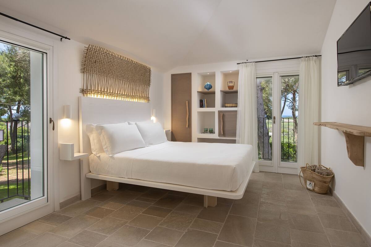 Un dormitorio con cama, mesita de noche, televisor montado en la pared y ventana con cortinas.