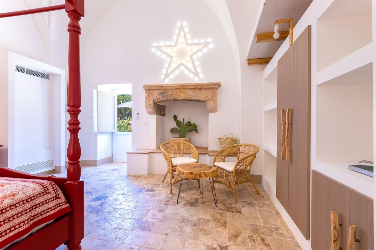 Un dormitorio con una cama con dosel, una chimenea iluminada por las estrellas, sillones de mimbre y suelo de piedra.