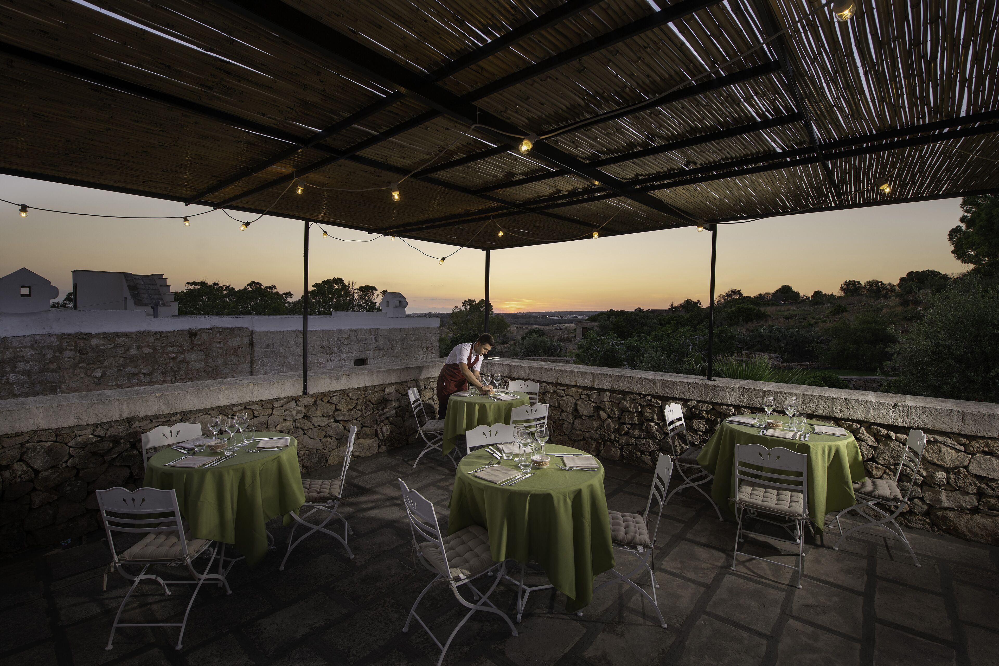 An outdoor dining area with tables set for two, covered by a roof, and overlooking a scenic landscape.