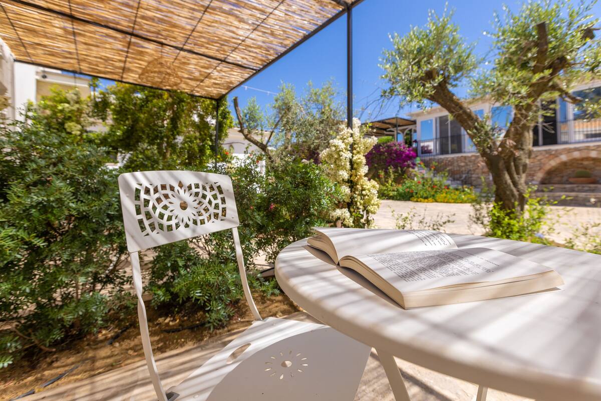 Un área de estar al aire libre con una mesa y una silla, rodeada de follaje y un edificio al fondo.