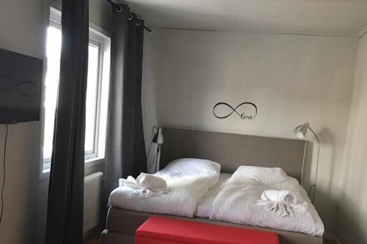 Quarto com cama, um banquinho vermelho, janela com cortinas, TV fixada na parede e luminária no teto.