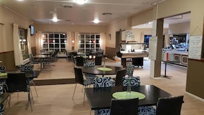 A spacious dining area with multiple tables and chairs, a kitchen area with a counter and appliances, and a television mounted on the wall. - Motoristen (Vara)
