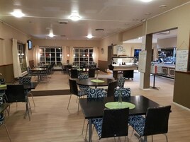 Uma sala de jantar ampla com várias mesas e cadeiras, uma cozinha com bancada e eletrodomésticos, e uma televisão fixada na parede.
