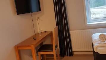 Huoneessa on puinen työpöytä, tuoli, valkoisella peitolla varustettu sänky, seinälle asennettu televisio ja ikkuna, josta avautuu näkymä puihin.