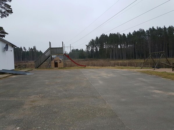 Un terrain de jeux avec un toboggan et des balançoires, un petit bâtiment et, au fond, une zone boisée.