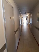 Corredor de hotel comprido e estreito, com portas em ambos os lados, piso ladrilhado e luminária no teto.