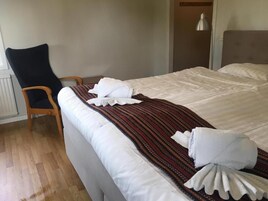 Uma cama bem arrumada com lençóis brancos e manta listrada, uma cadeira de madeira e uma mesa de cabeceira com abajur.