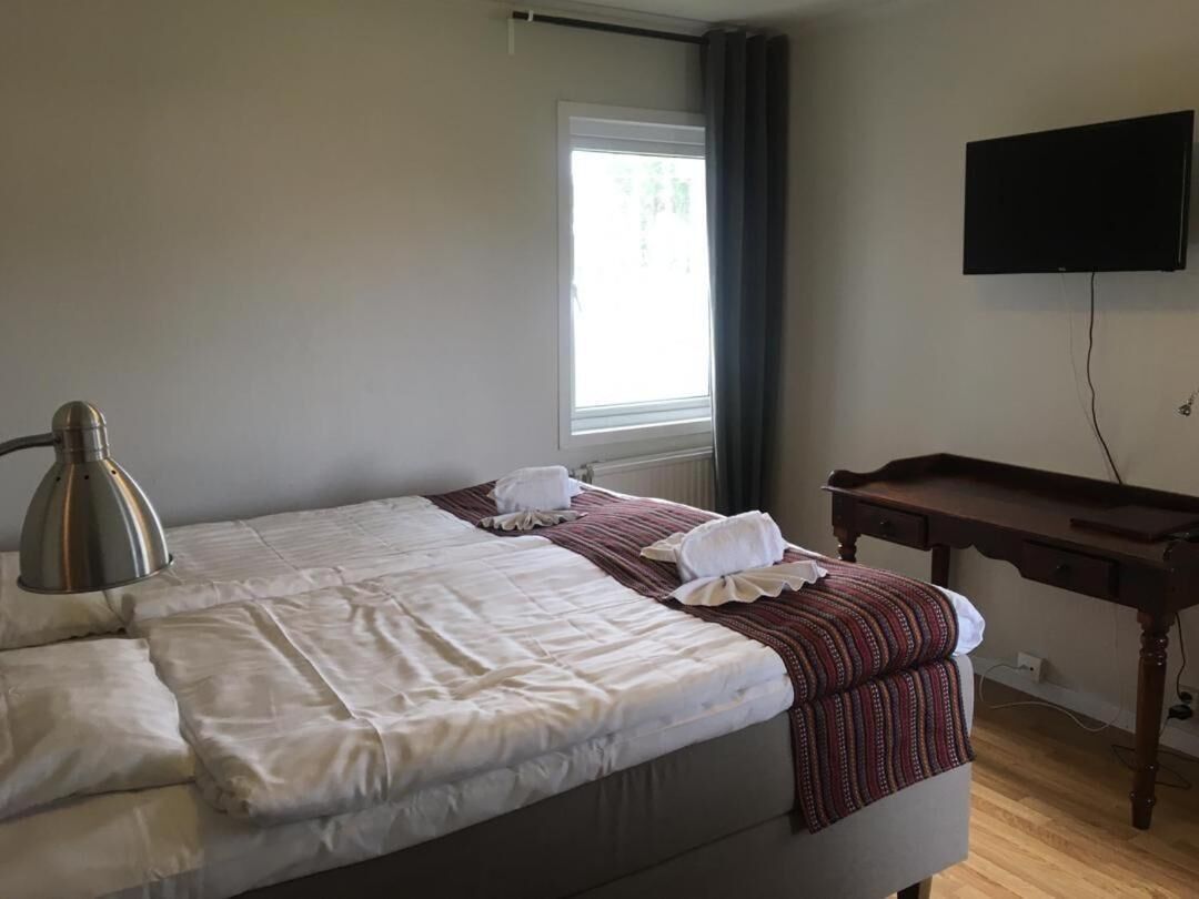 Cama bem arrumada com lençóis brancos, luminária de cabeceira e janela com cortinas. Uma TV de tela plana está montada na parede à direita.