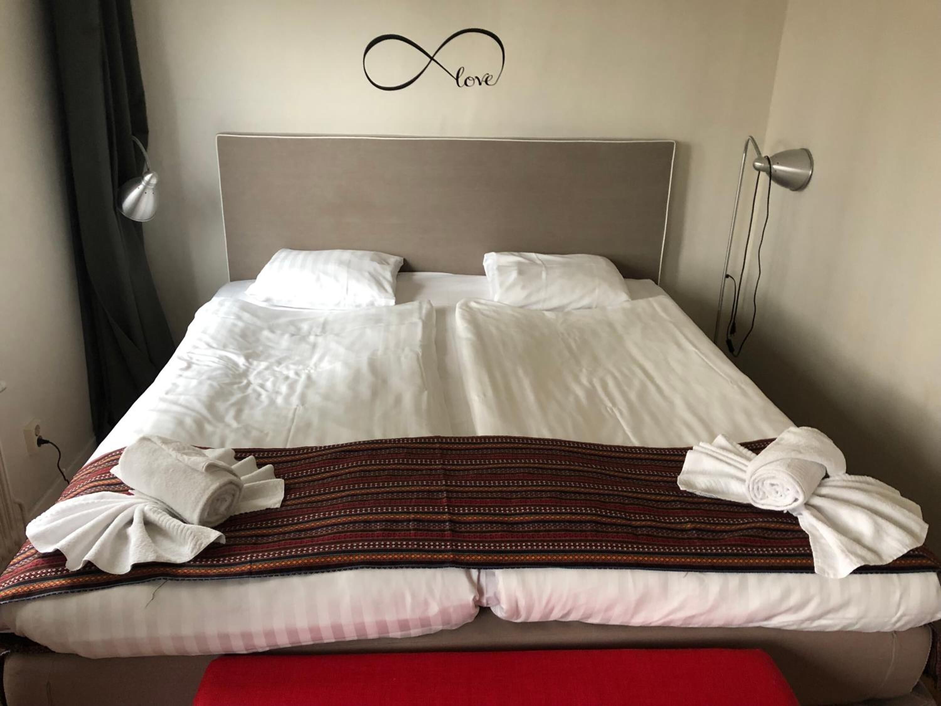 Un lit bien fait, avec du linge de lit blanc, un symbole de l’infini décoratif au-dessus, et un banc rouge au pied du lit.