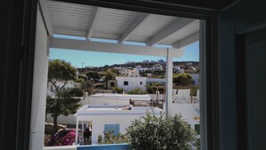 A view from a window overlooking a hillside with white buildings, a balcony with a table and chairs, and a garden with pink flowers. - Angelos Rooms (Naxos)