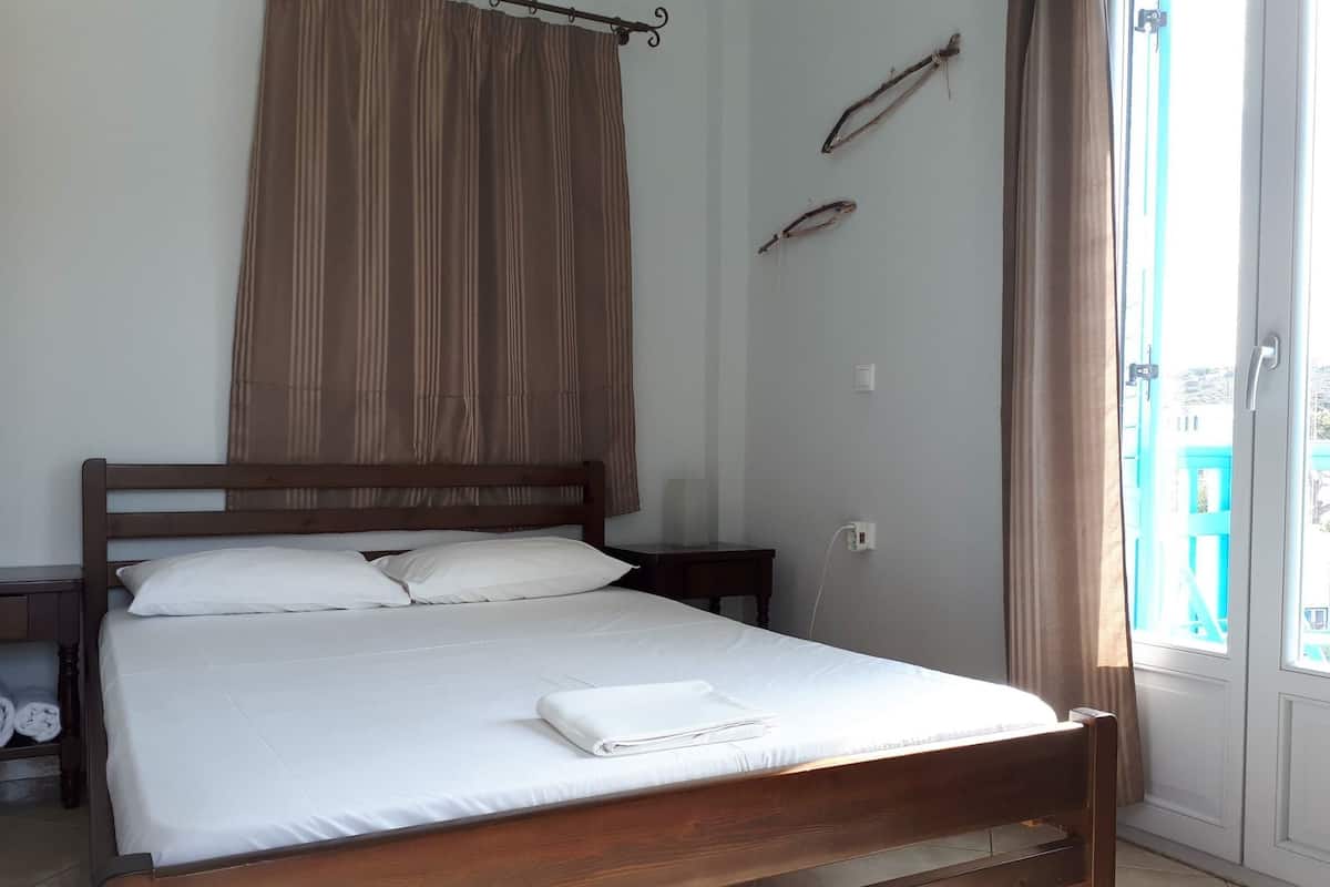 A bedroom with a wooden bed, white bedding, a curtain, a nightstand, and a view of the outside.