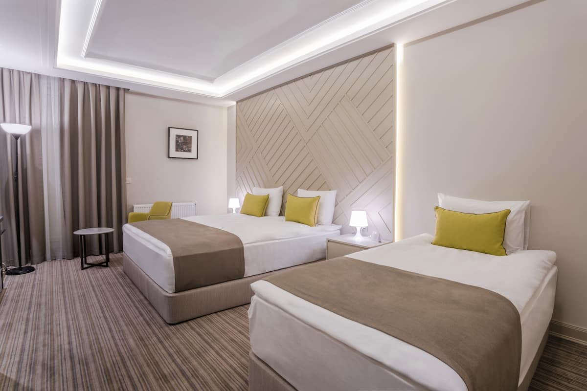 A hotel room with two beds, a small table, a lamp, and a radiator.