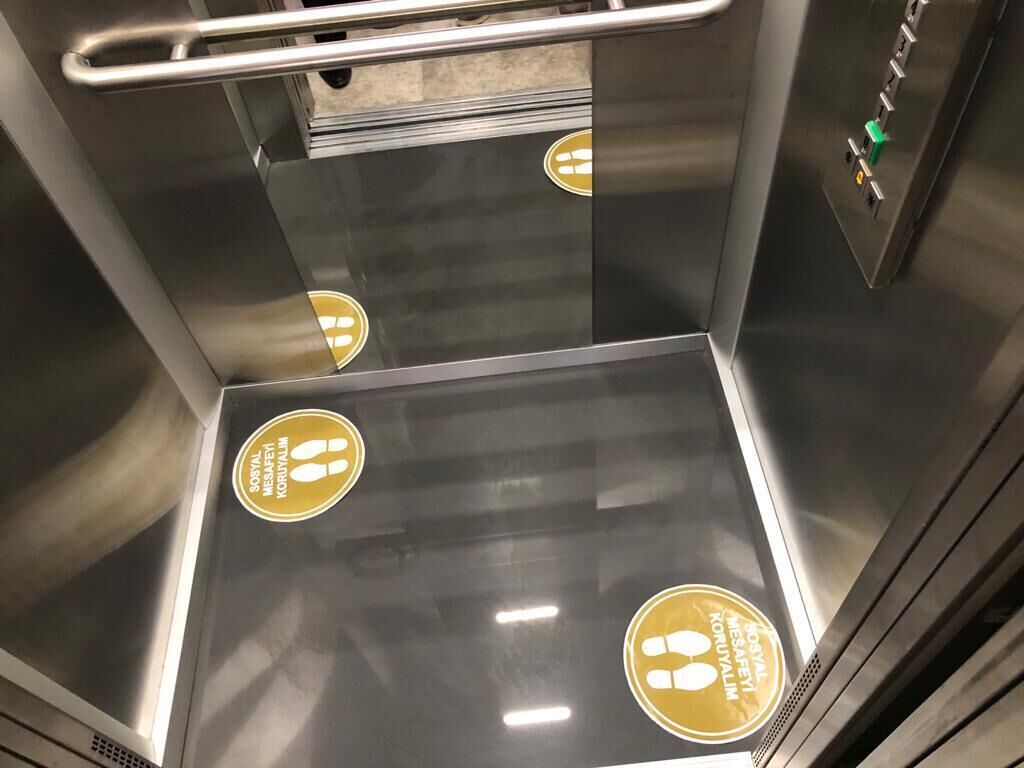 L’intérieur d’un ascenseur, avec des parois métalliques et un sol réfléchissant, agrémenté de symboles de sécurité et d’une barre d’appui.