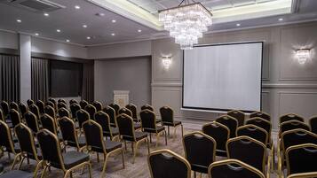 Un salón de conferencias con filas de sillas negras de marco dorado, una pantalla grande y un candelabro.