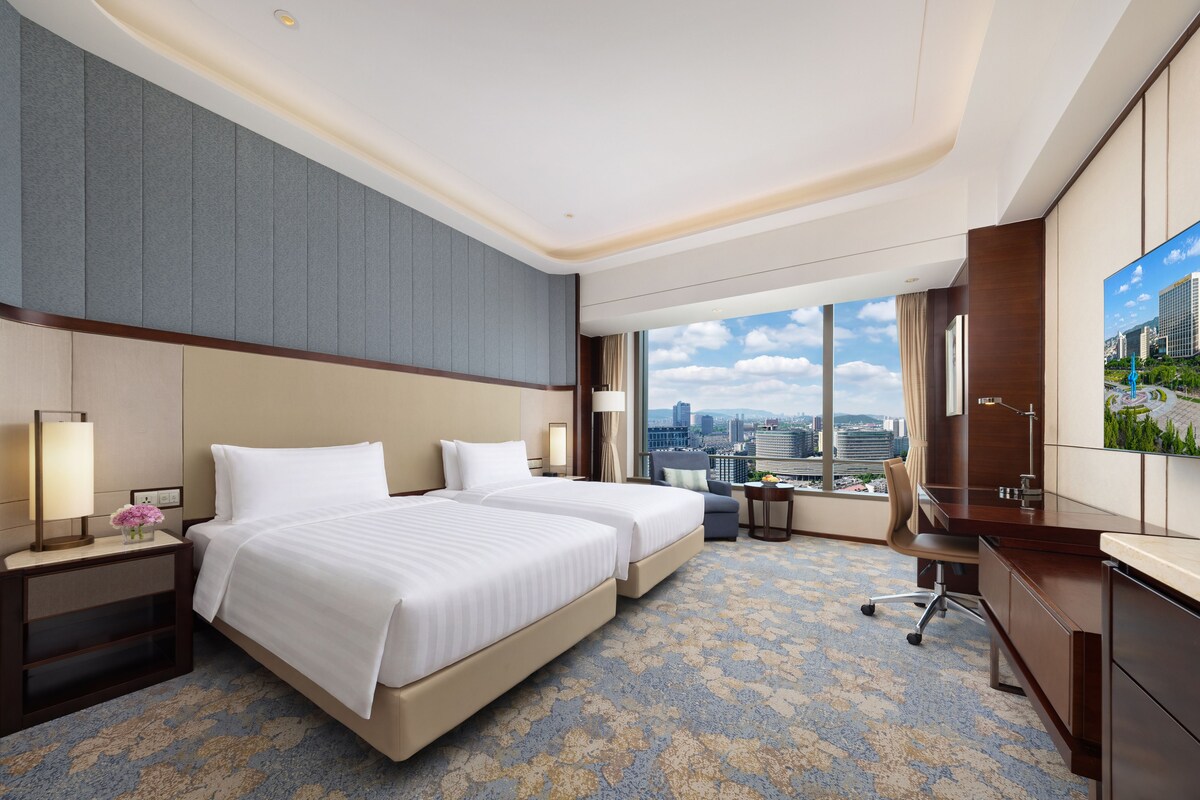 Habitación de hotel con dos camas, un escritorio, una silla, un televisor y vistas a la ciudad.