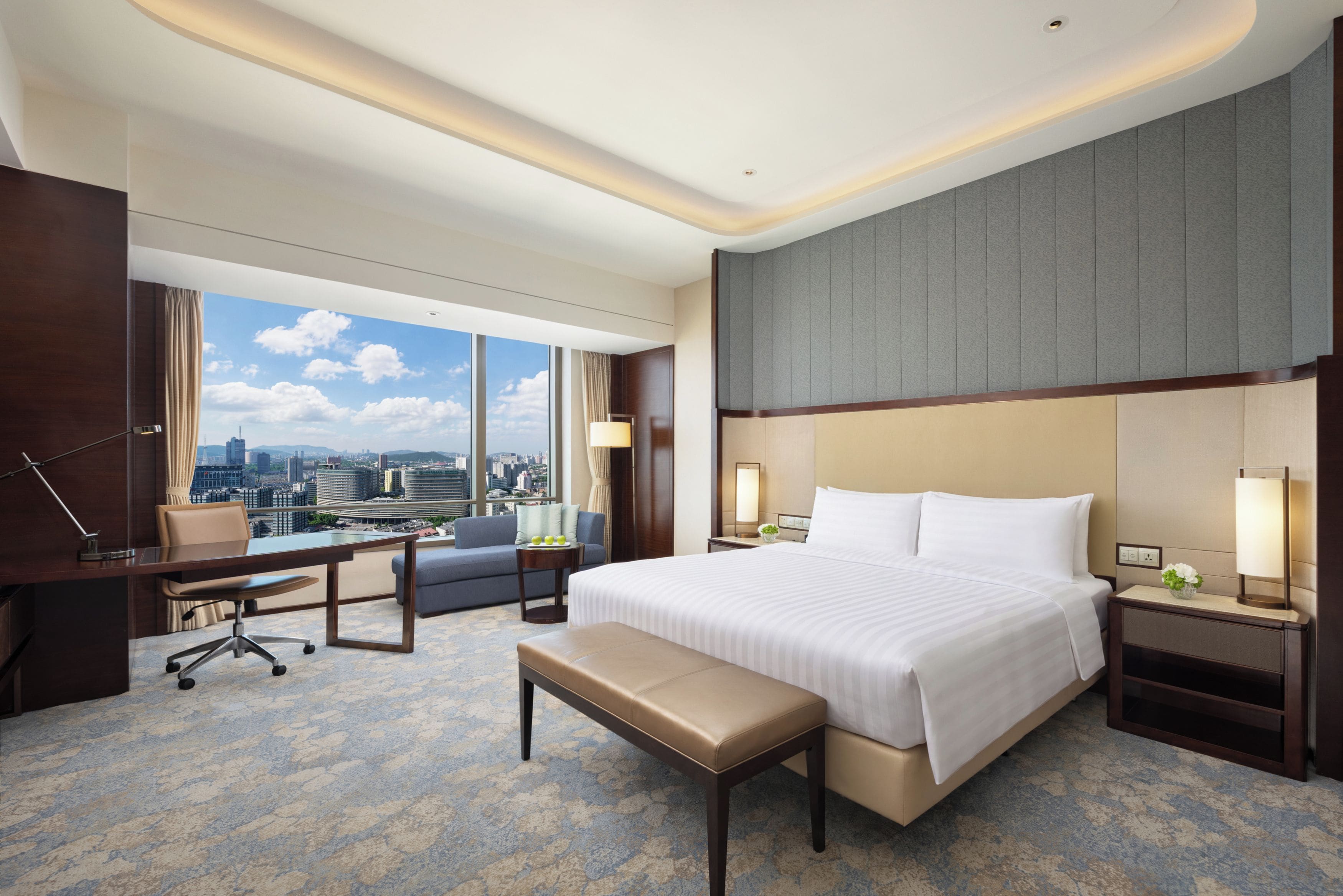Habitación de hotel con una cama grande, un escritorio, una silla, un banco y vistas a un paisaje urbano a través de amplios ventanales.