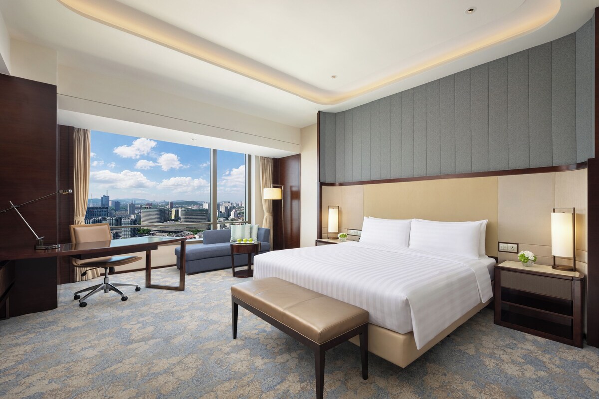Habitación de hotel con una cama grande, un escritorio, una silla, un banco, una mesita de noche y vistas a la ciudad.