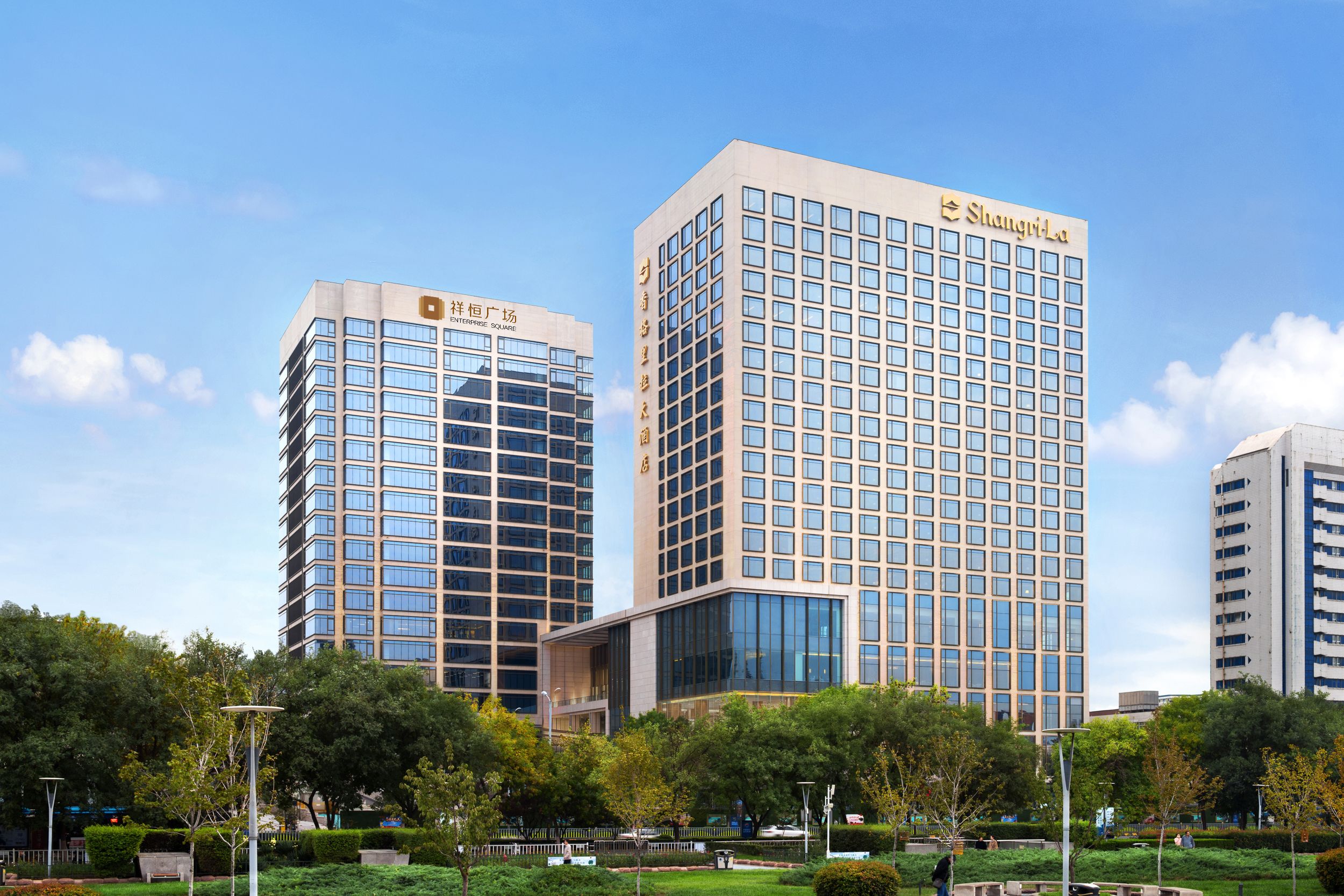 Two high-rise buildings with commercial signage, surrounded by greenery and a clear sky.