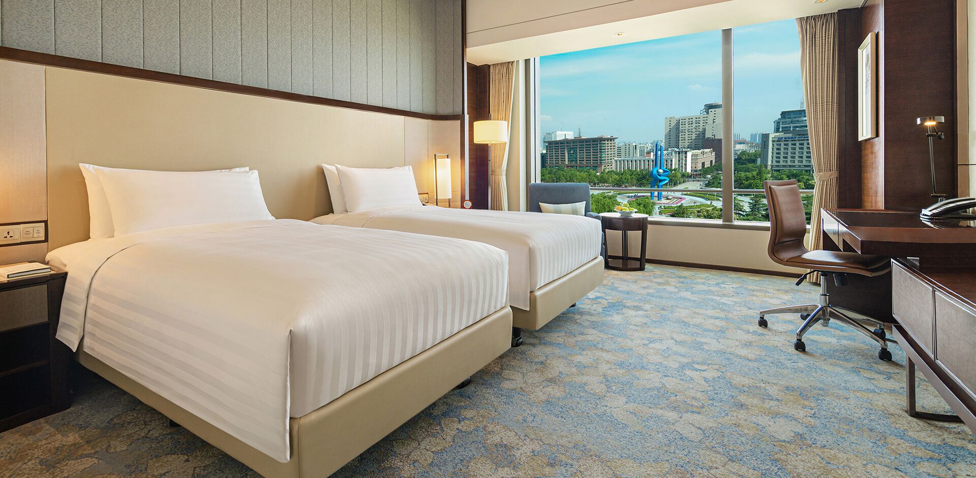 A hotel room with two beds, a desk, a chair, and a view of the cityscape.
