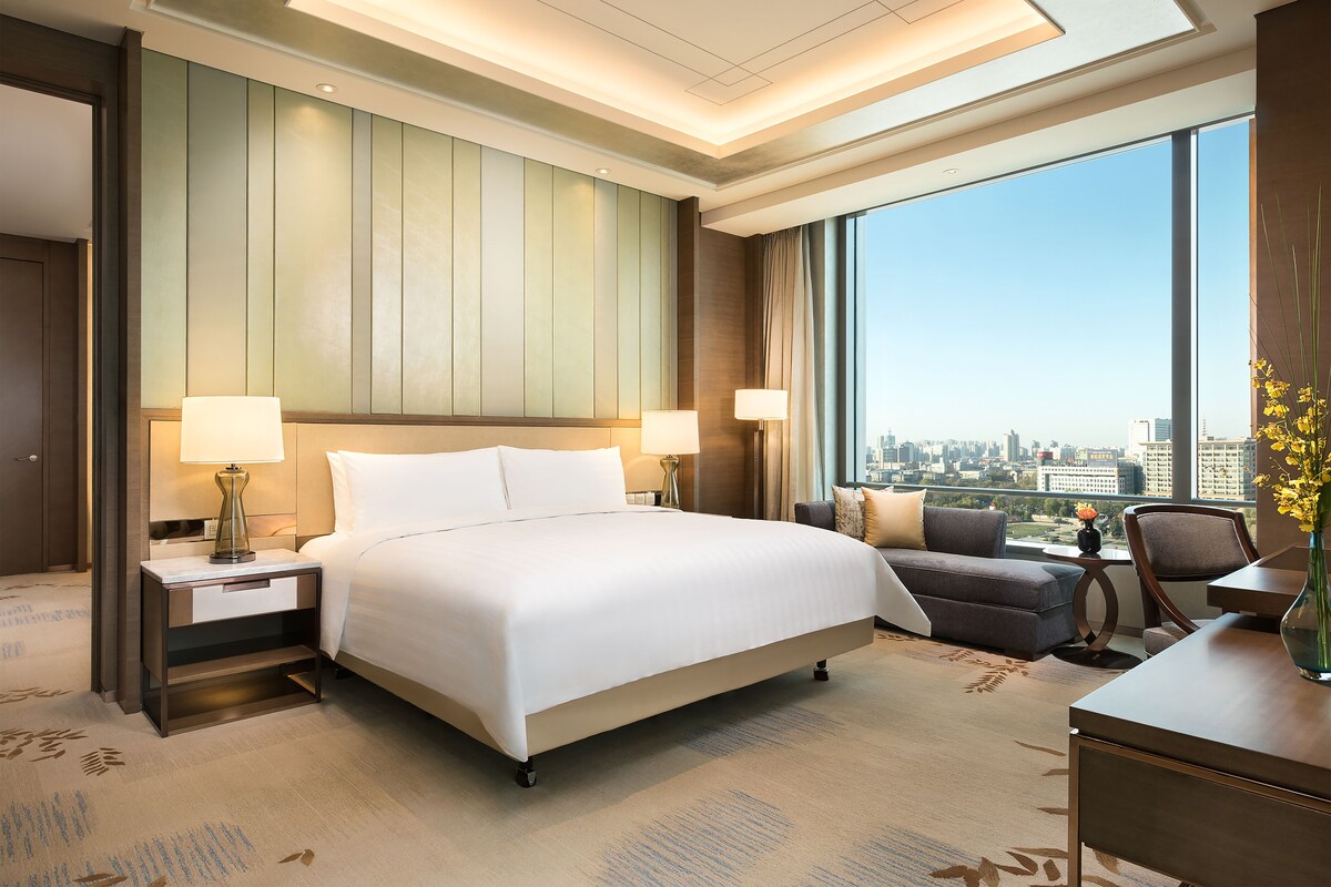 Habitación de hotel con una cama grande, dos mesitas de noche con lámparas, un escritorio con silla y vistas a la ciudad a través de amplios ventanales.