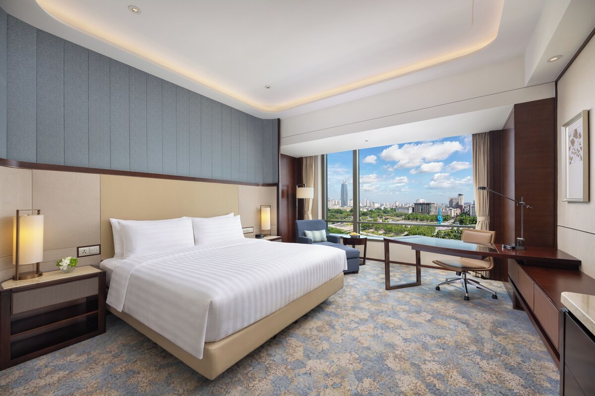 Habitación de hotel con una cama grande, un escritorio y vistas a un paisaje urbano.