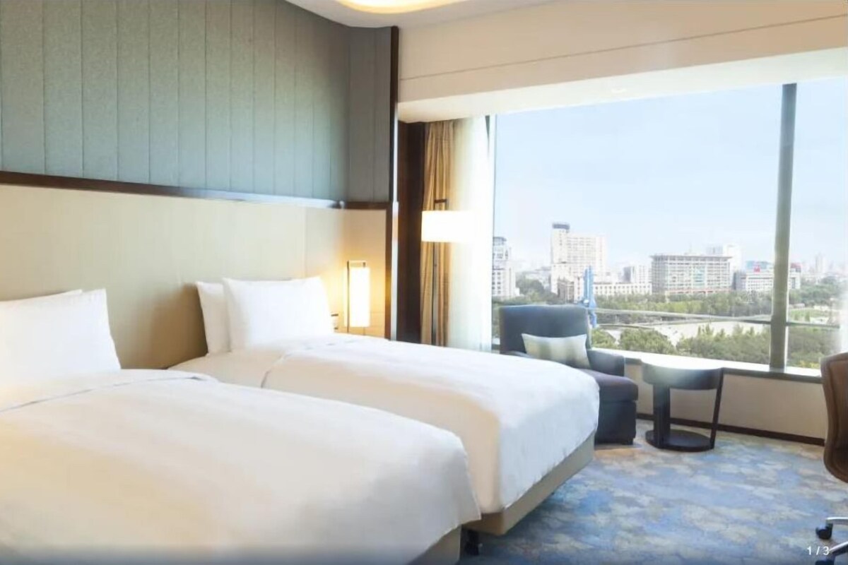 Habitación de hotel con dos camas, un ventanal grande con vista a la ciudad, una mesita y una silla.
