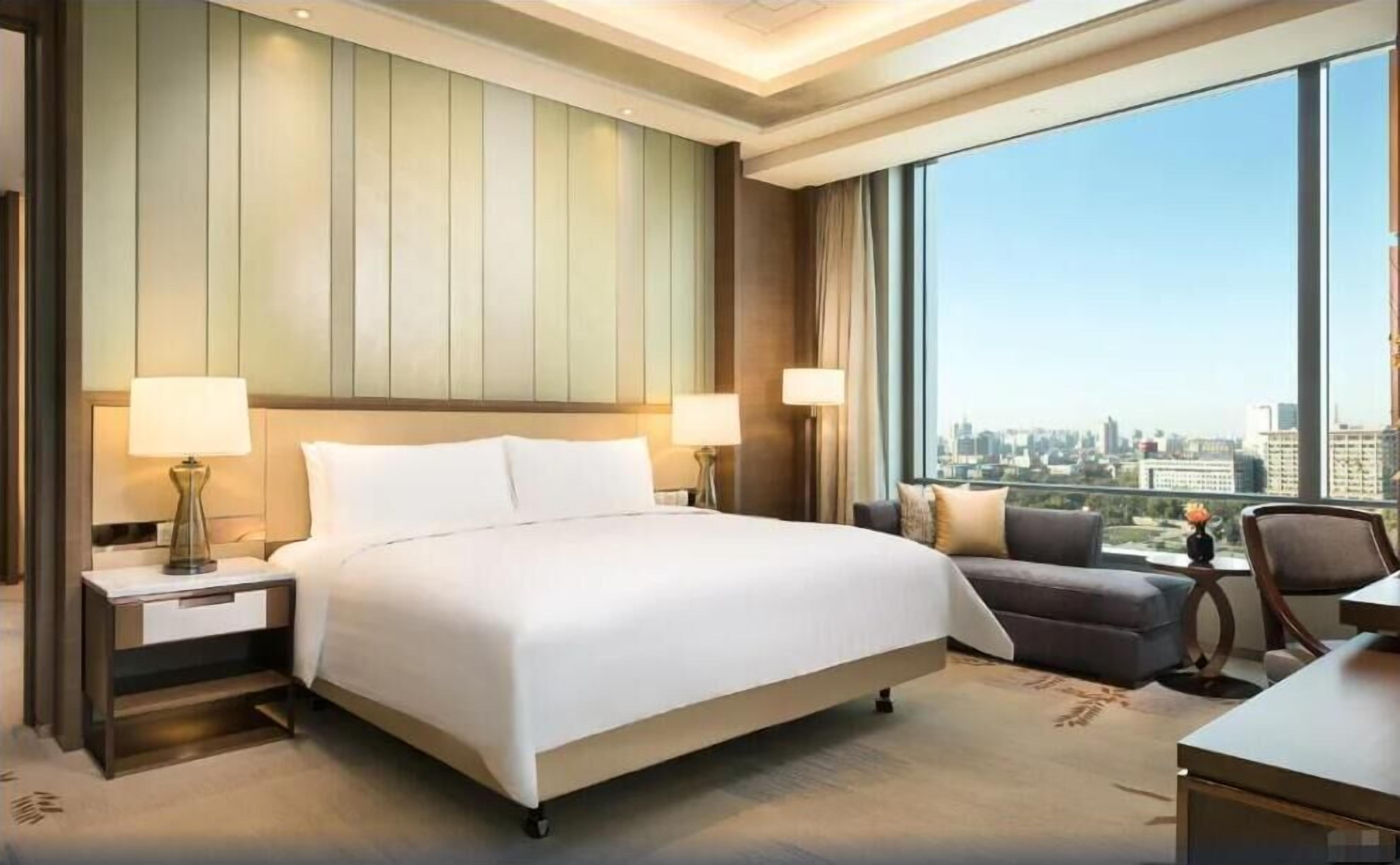 Een hotelkamer met een groot bed, twee nachtkastjes met lampen, een bank en uitzicht op de stad.