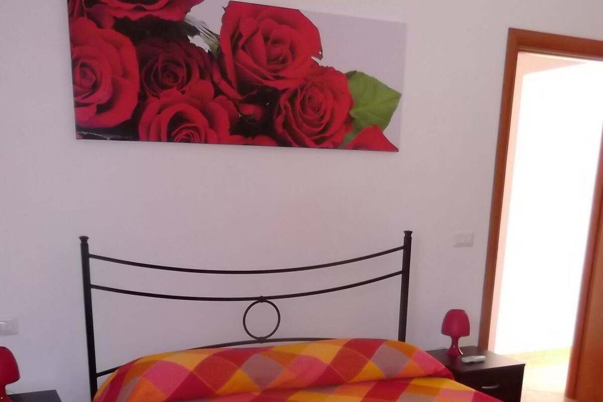Un dormitorio con cama, mesita de noche, lámpara y un cuadro de rosas rojas en la pared.