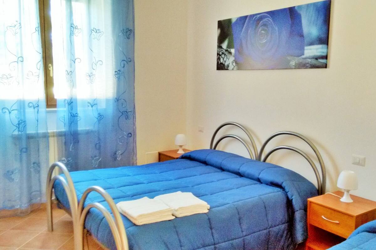 Un dormitorio con cama, dos mesitas de noche, un velador, una ventana con cortinas y un cuadro en la pared.