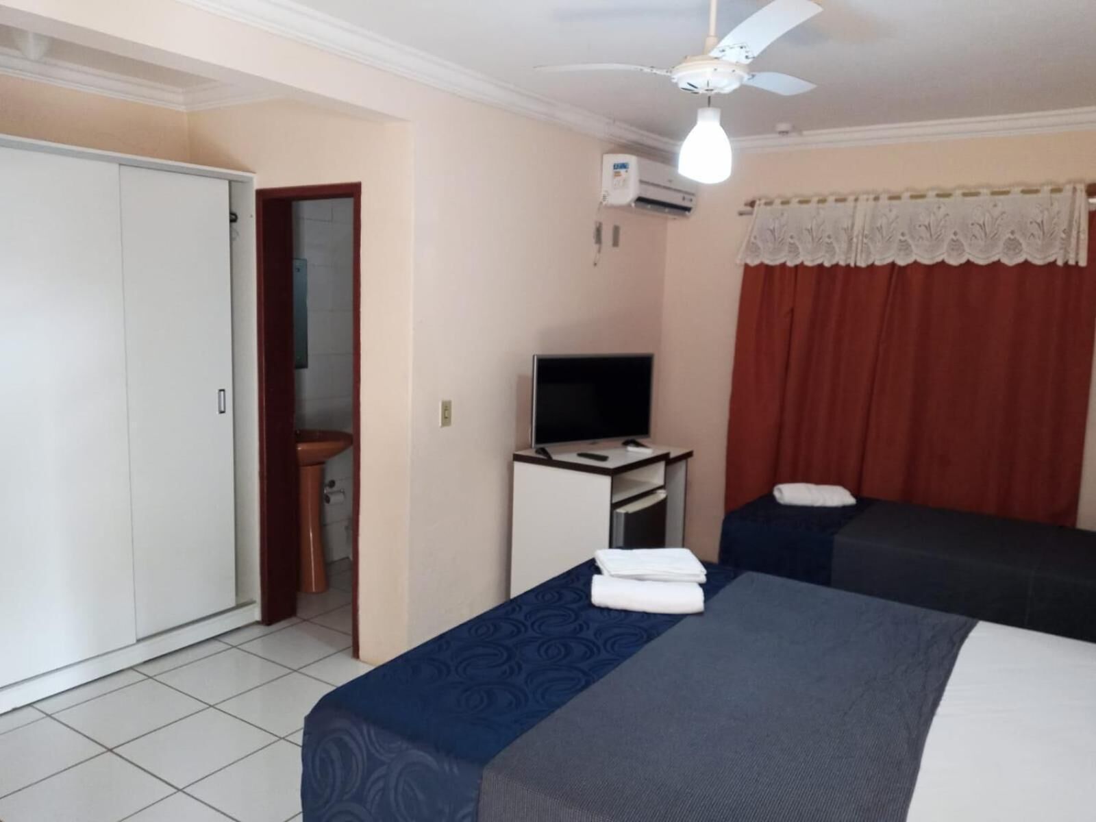A hotel room with a bed, a desk with a TV, a wardrobe, and a ceiling fan.