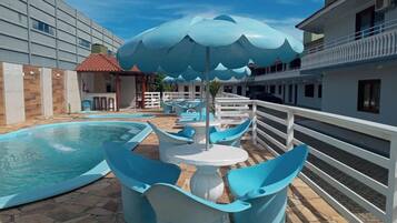 A swimming pool area with blue chairs and umbrellas, a stone-tiled deck, and a building with balconies.