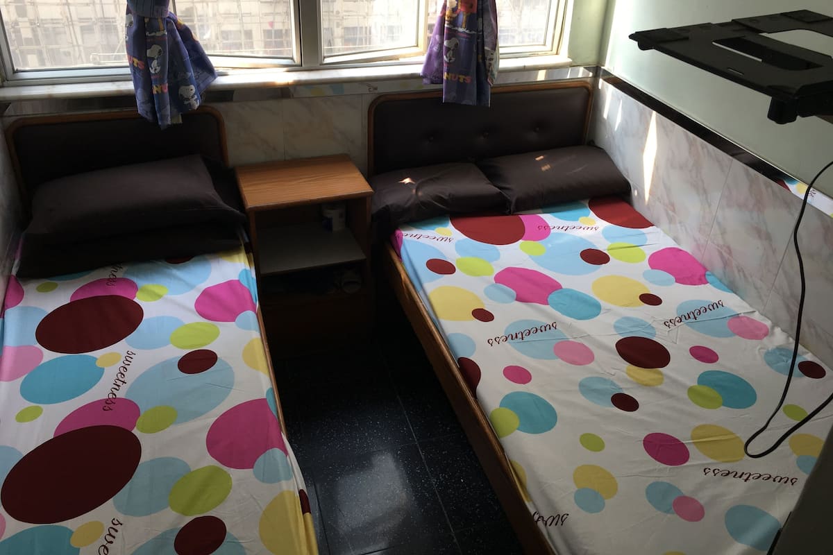 A small, compact room with two single beds, a small wooden table, and a window with curtains.