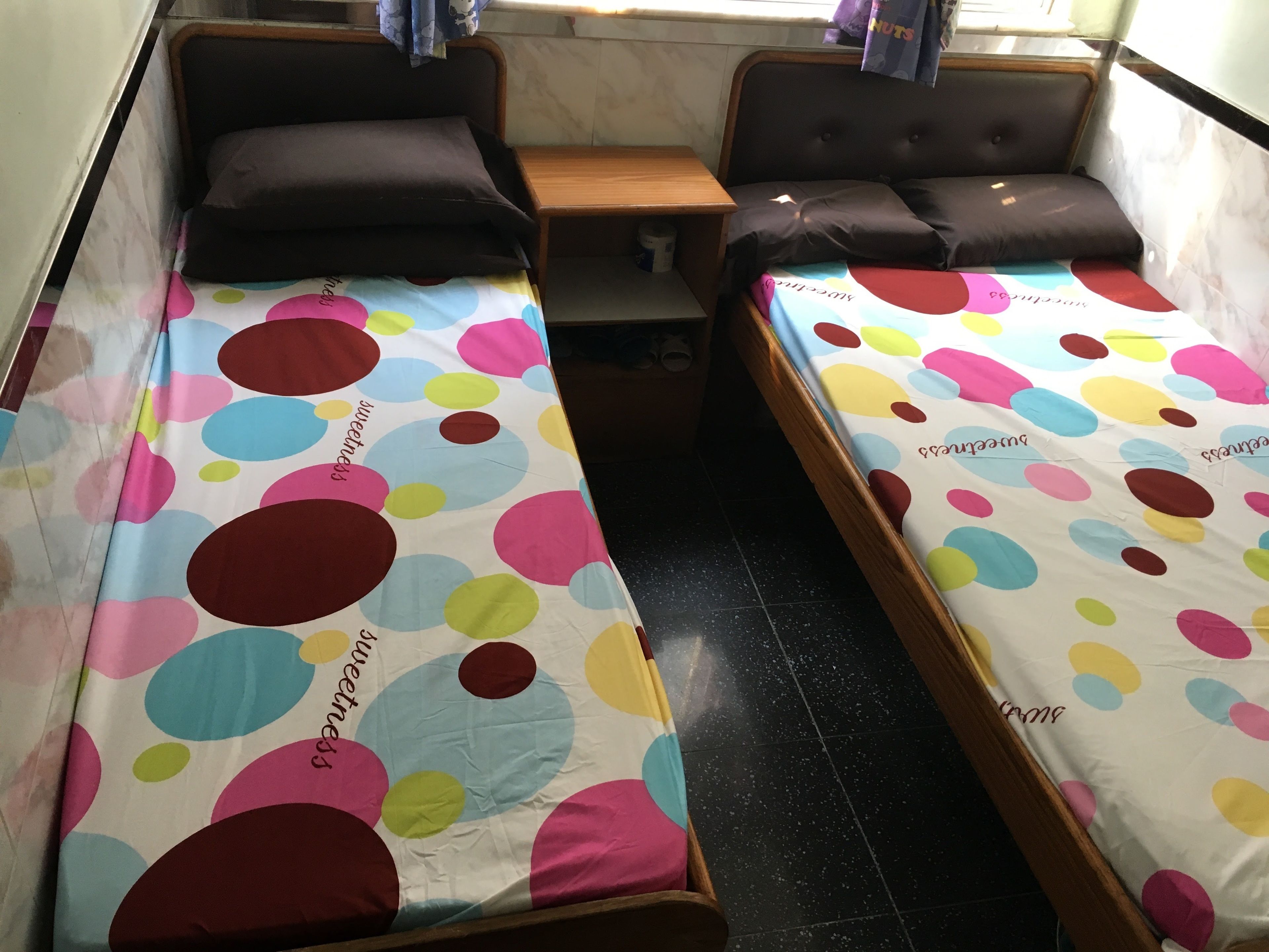 A room with two single beds, each with a polka dot bedspread. There is a wooden nightstand between the beds, and a small table with a chair. The room has a window with curtains, and the walls are tiled.