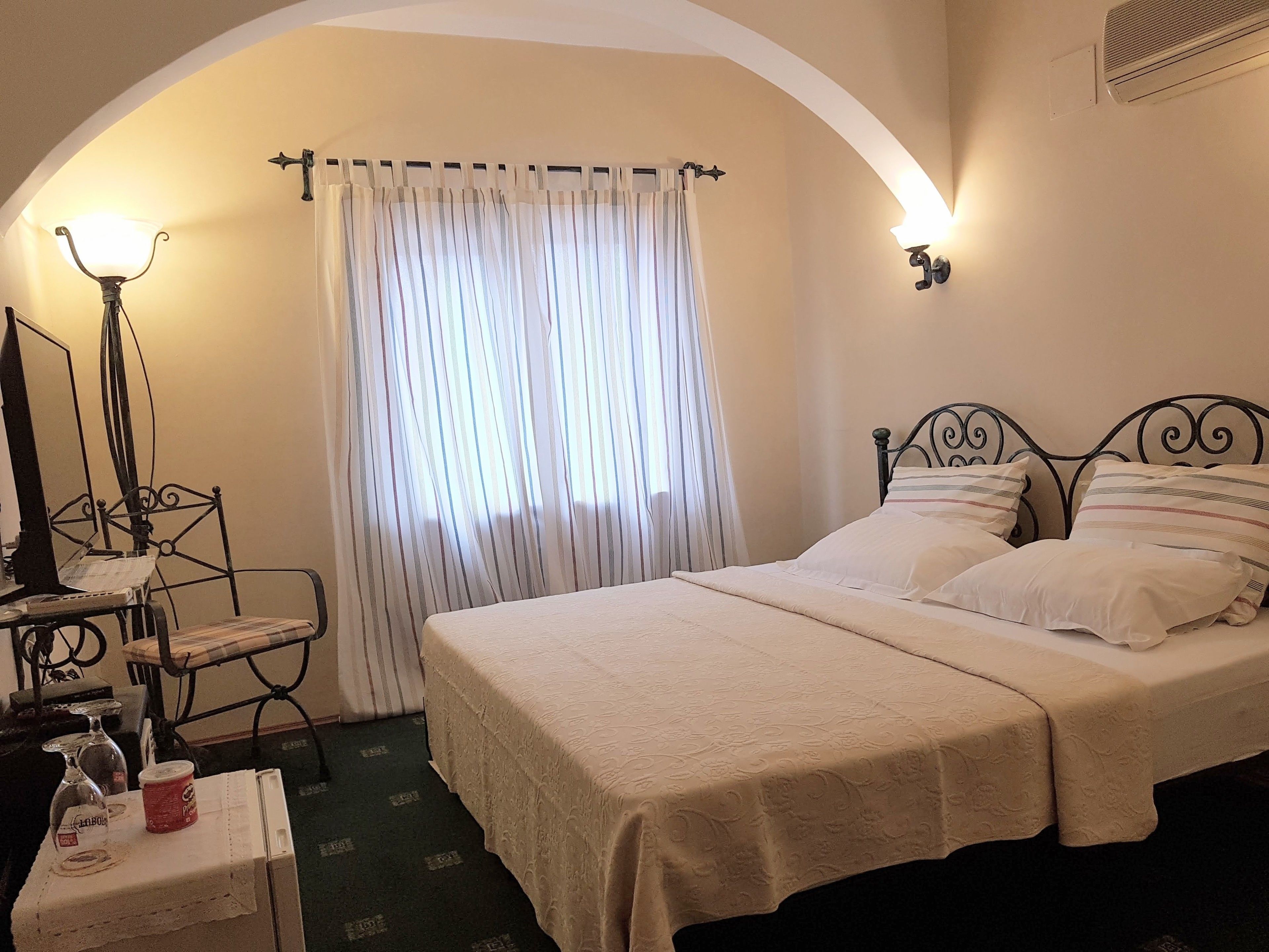 Quarto com cama, escrivaninha com computador, luminária e janela com cortinas.