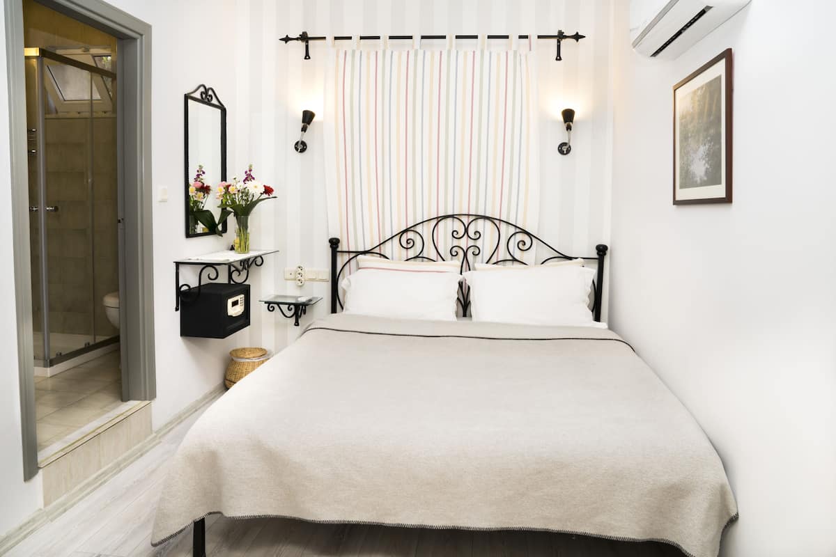 A bedroom with a bed, a bedside table, a mirror, a wall-mounted shelf with flowers, and a window with curtains.