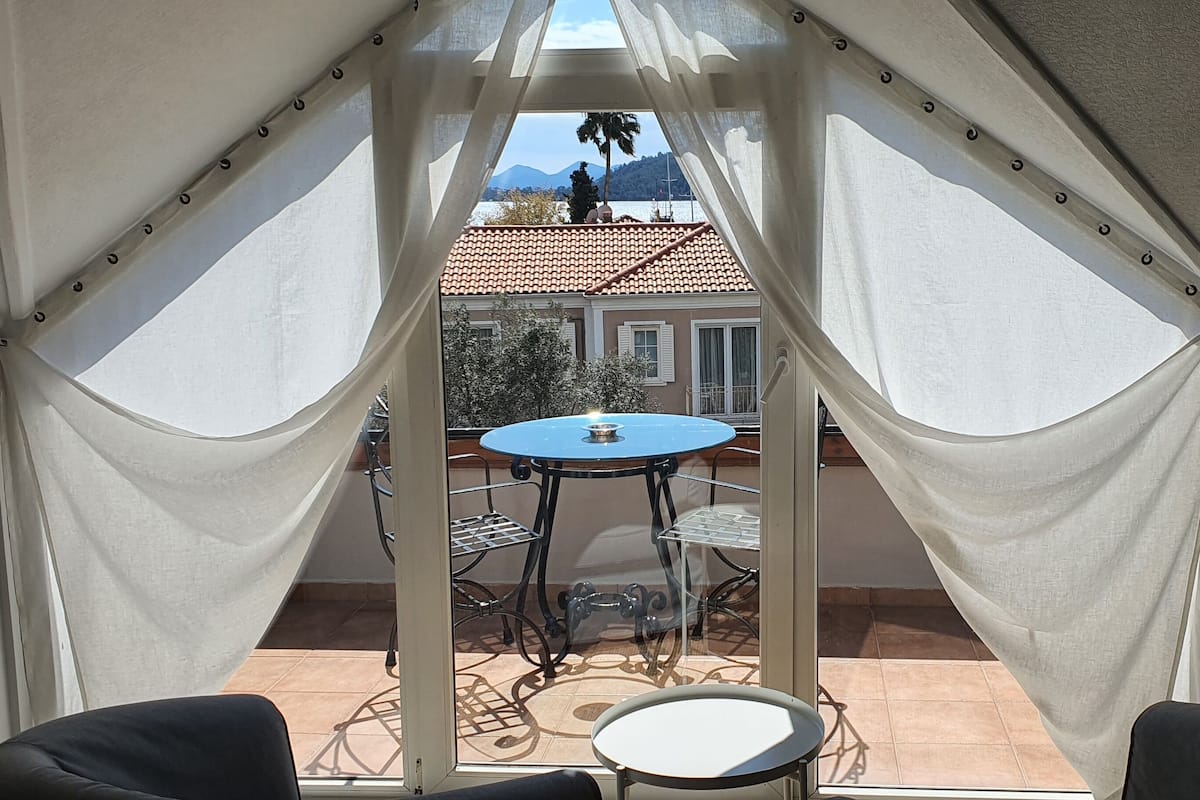A room with a view of a patio area, featuring a table and chairs, and a balcony with a railing.