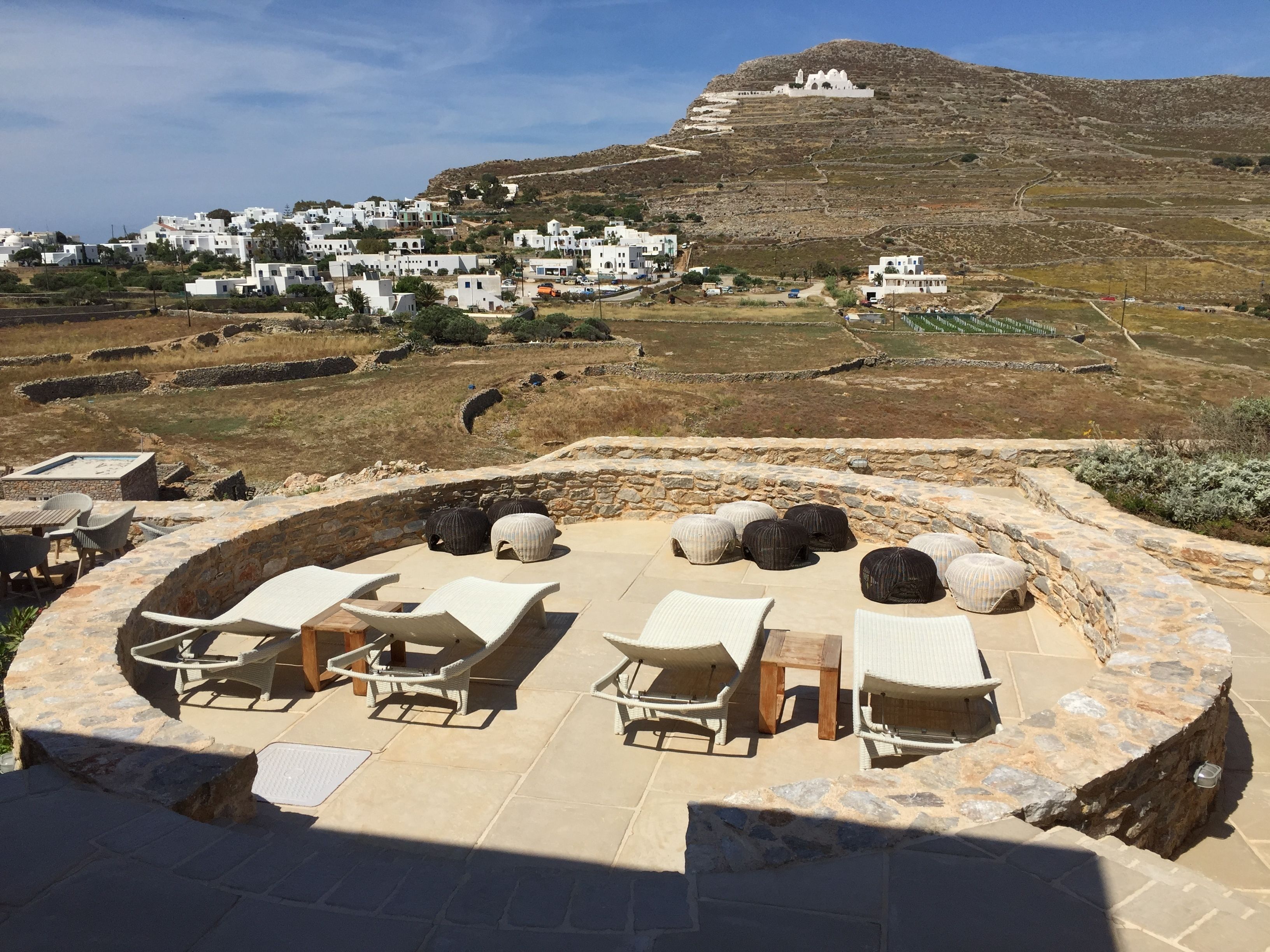 A sunlit outdoor seating area with stone walls and lounge chairs, overlooking a hillside village with white buildings.