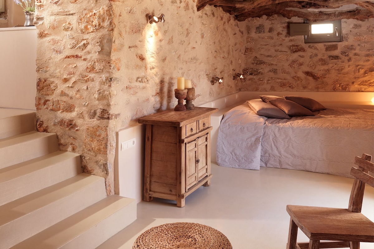 A rustic bedroom with exposed stone walls, a wooden bed, and a small wooden chest of drawers.