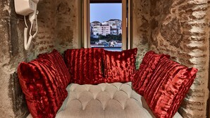 Quarto com cama, almofadas vermelhas, telefone branco e janela com vista para edifícios.