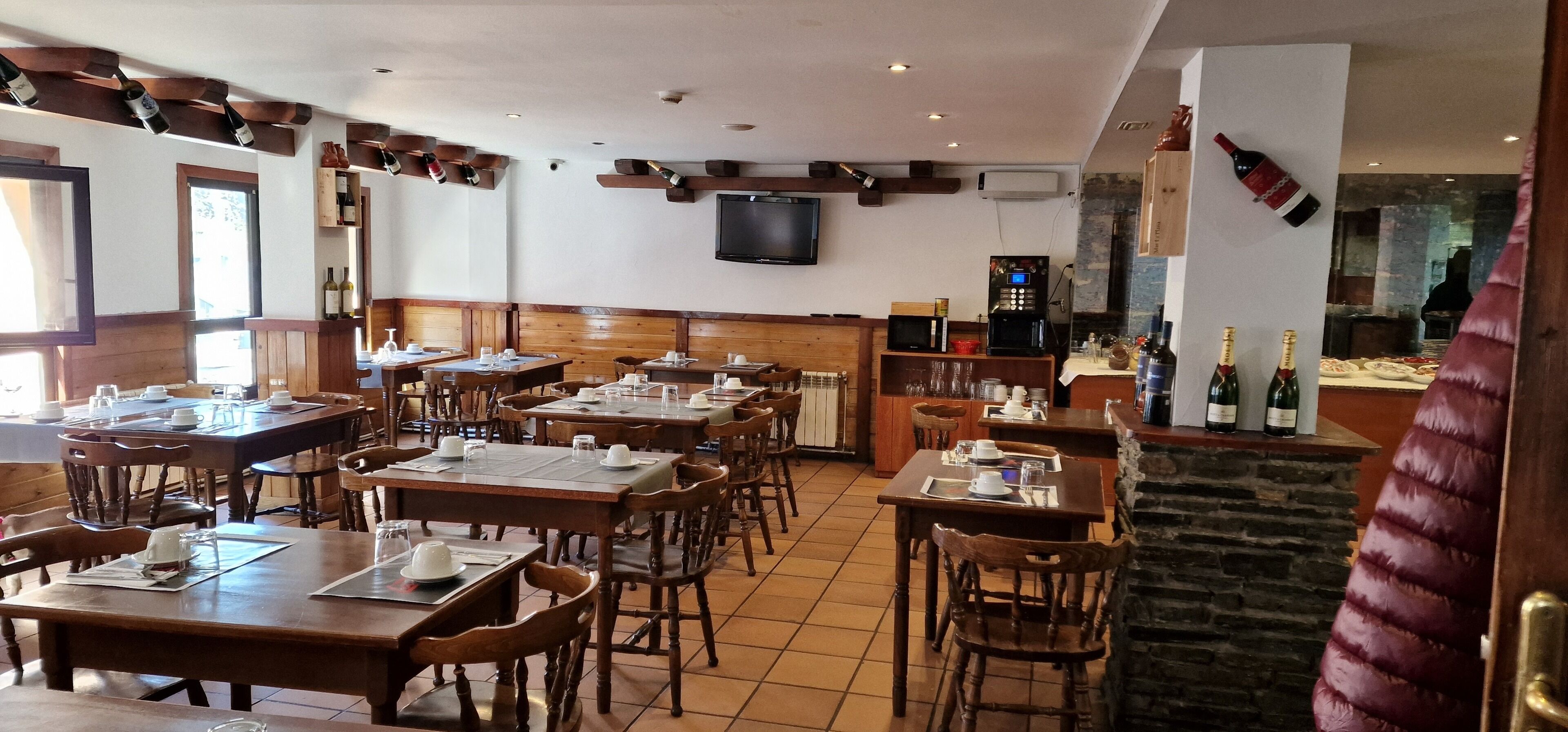 A restaurant with wooden tables and chairs, a TV mounted on the wall, and a bar area with bottles.
