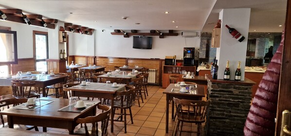 Un restaurante con mesas y sillas de madera, un televisor montado en la pared y una zona de bar con botellas.