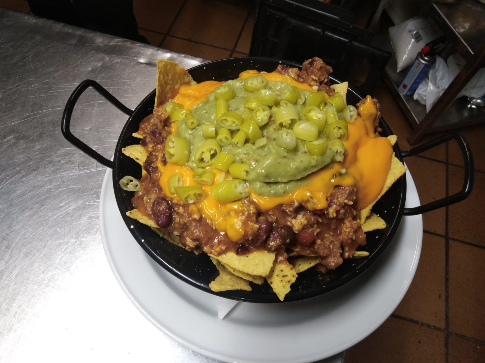 A black skillet filled with nachos topped with cheese, jalapeños, and ground meat, placed on a white plate on a metal table.