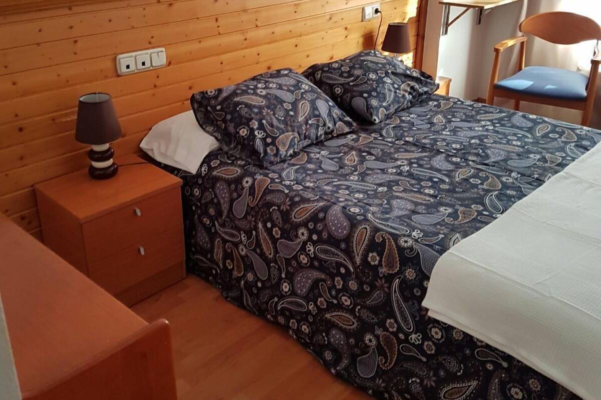 Een slaapkamer met een houten hoofdbord, een bed met beddengoed met een patroon, een nachtkastje met een lamp en een stoel.