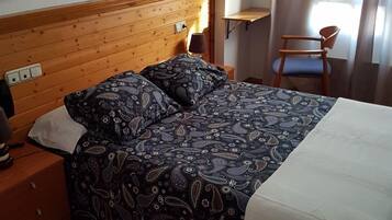 Habitación de hotel con cabecera de madera, cama con ropa de cama estampada, mesita de noche, lámpara, silla y ventana con cortinas.