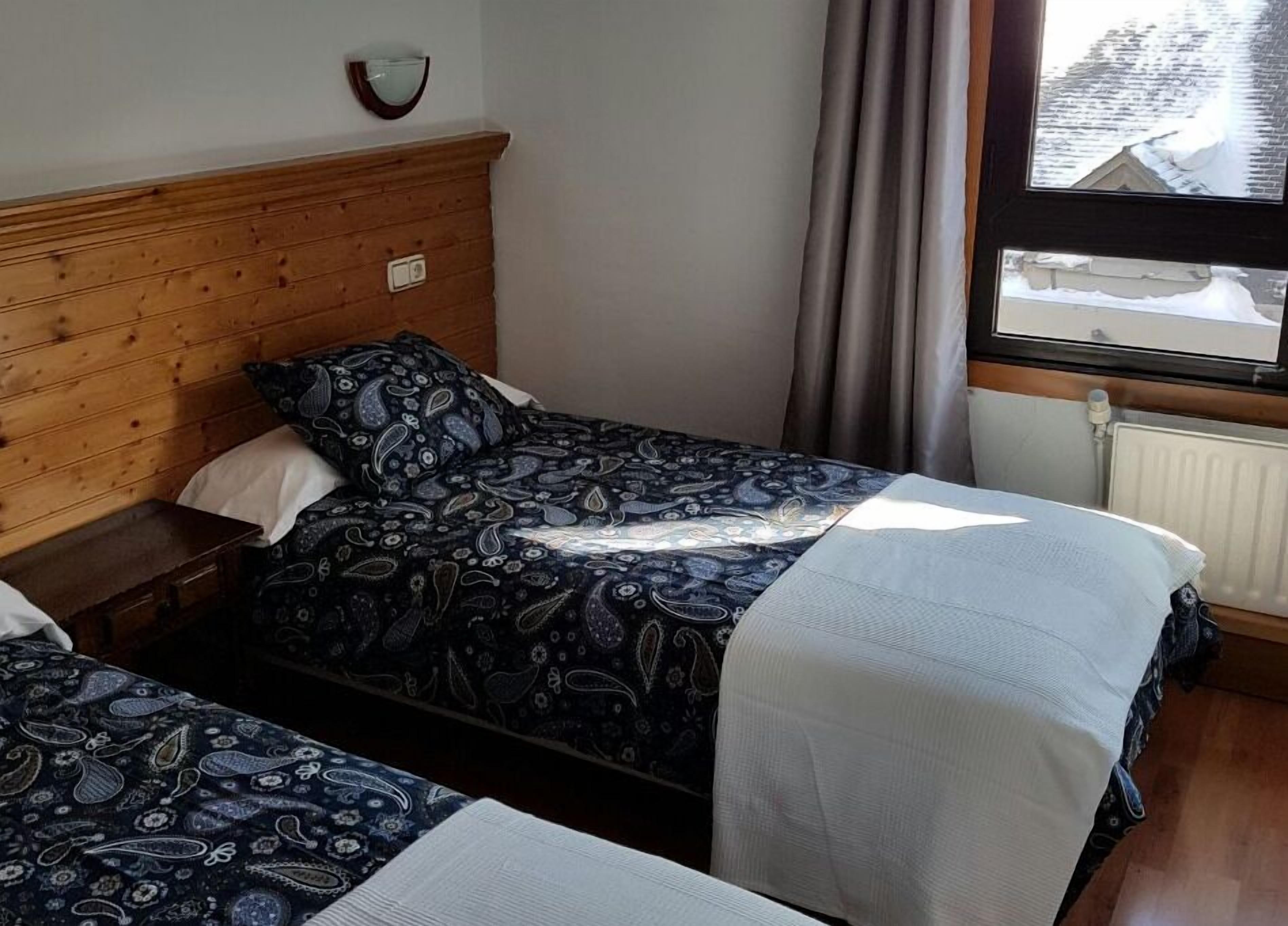 A room with two beds, wooden headboards, a window with a view of snow-covered roofs, and a wall-mounted light fixture.