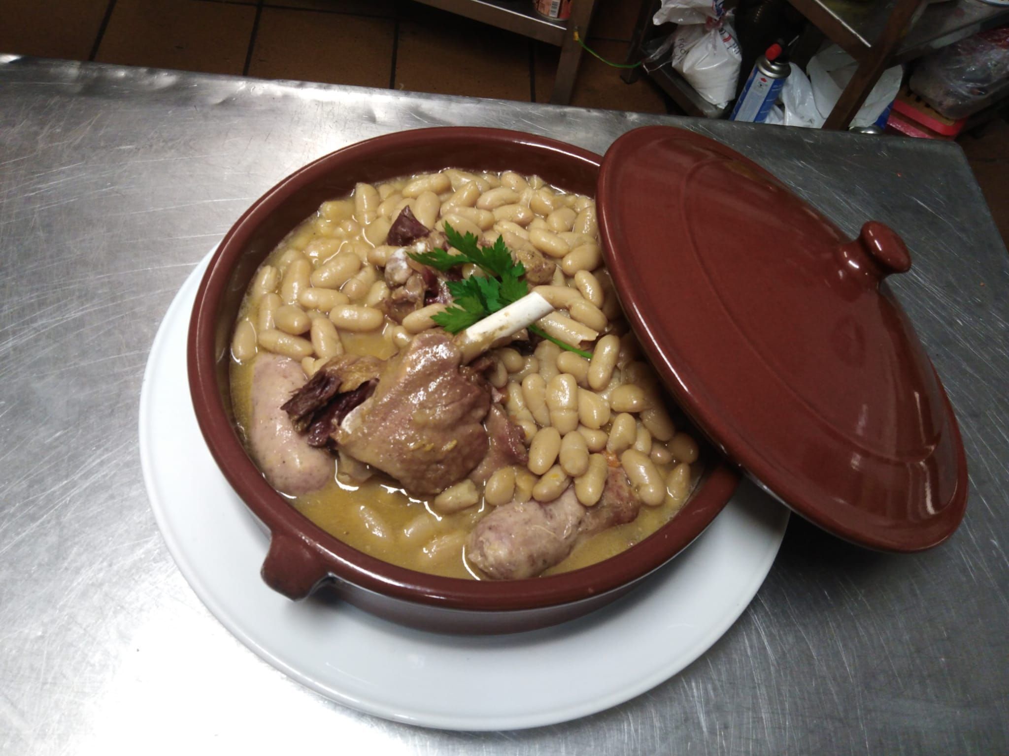 A large ceramic pot with a lid, containing a stew with beans and meat, placed on a metal table.