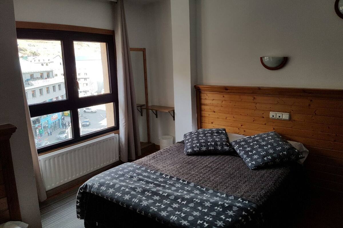 Een slaapkamer met een bed, twee kussens, een houten hoofdbord, een raam met uitzicht op gebouwen en een wandlamp.