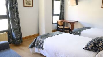 Habitación de hotel con dos camas, una mesa de madera, una silla y un ventanal con cortinas.